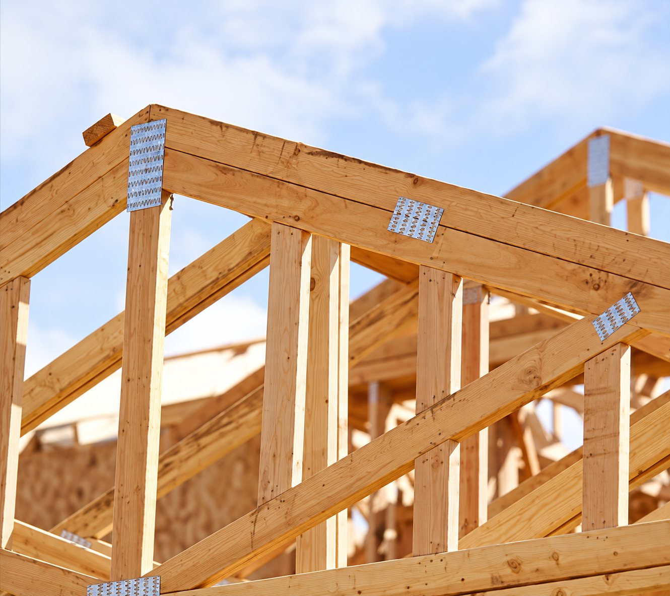 Structure de charpente en bois clair avec connecteurs métalliques sur une maison en cours de construction, vue en contre-plongée sous un ciel nuageux.