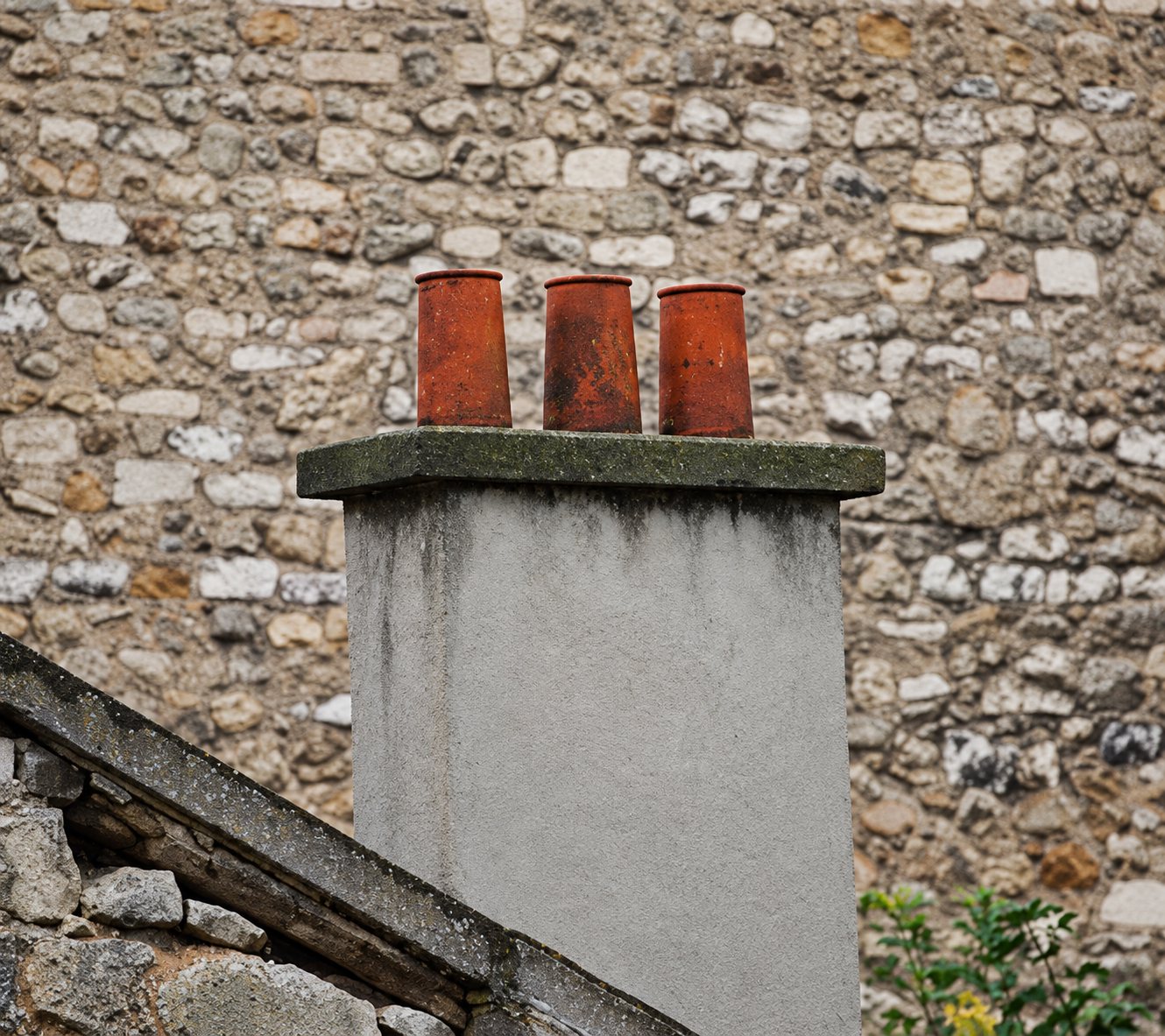Une cheminée grise surmontée de trois mitrons en terre cuite se dresse devant un vieux mur en pierres apparentes.
