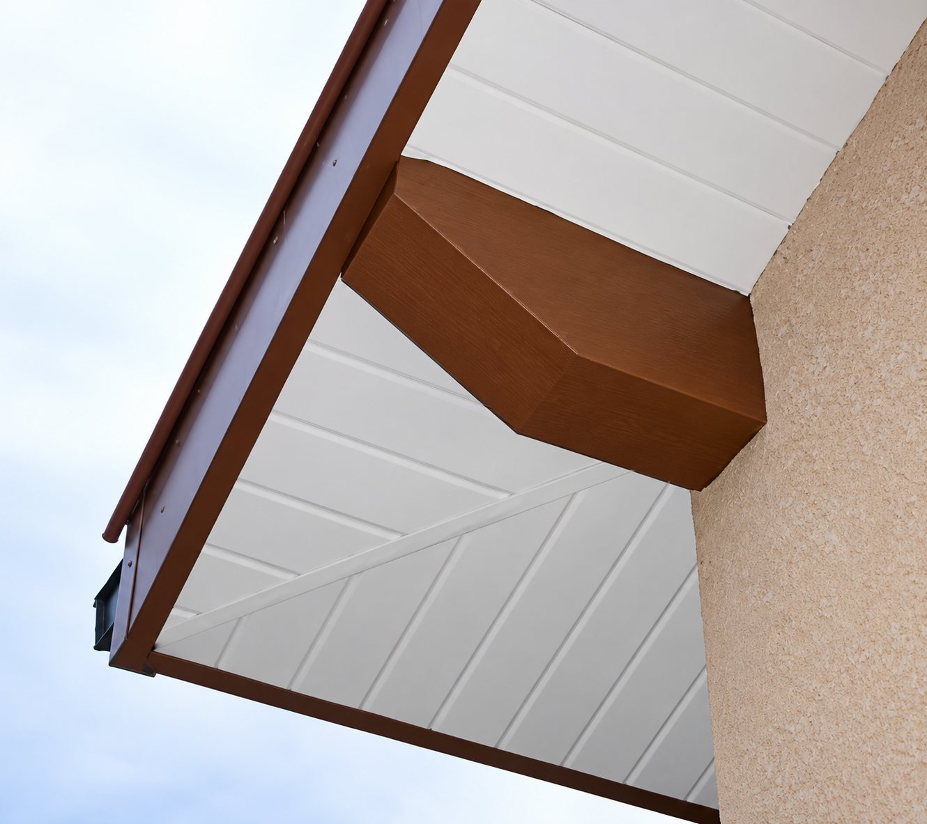 Vue en contre-plongée d'un débord de toit dont la sous-face en lambris blanc est bordée d'un habillage marron, contre un mur crépi beige.
