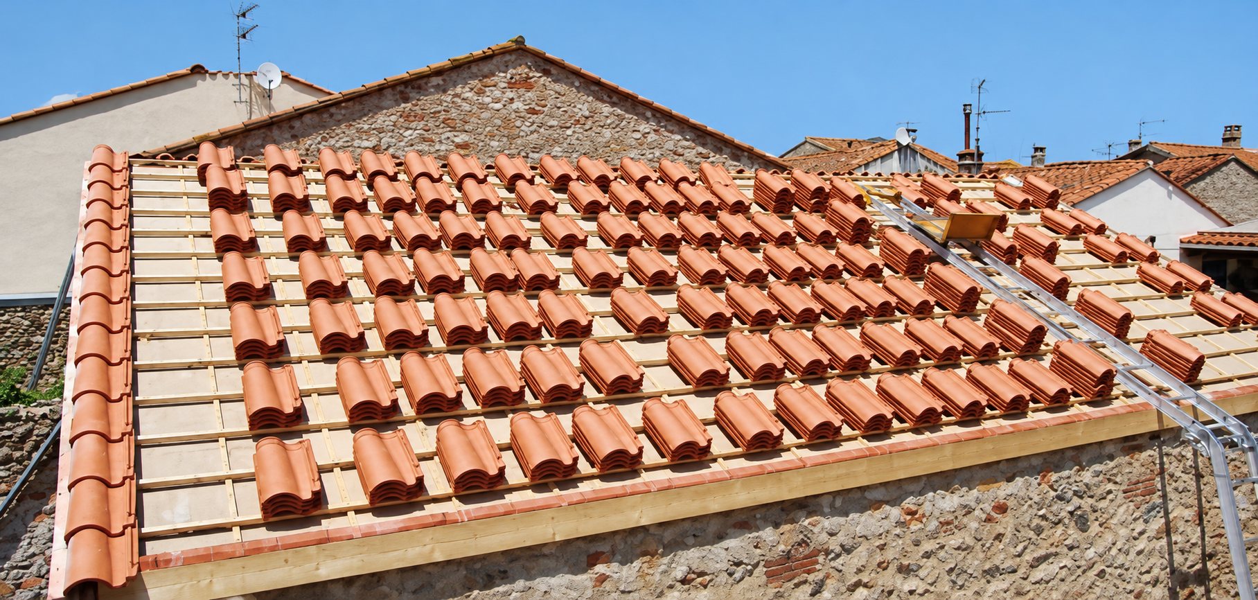 Toit en rénovation sur lequel des rangées de tuiles canal orangées sont posées sur un support clair, avec un monte-matériaux sur le côté et des maisons en pierre alentour.