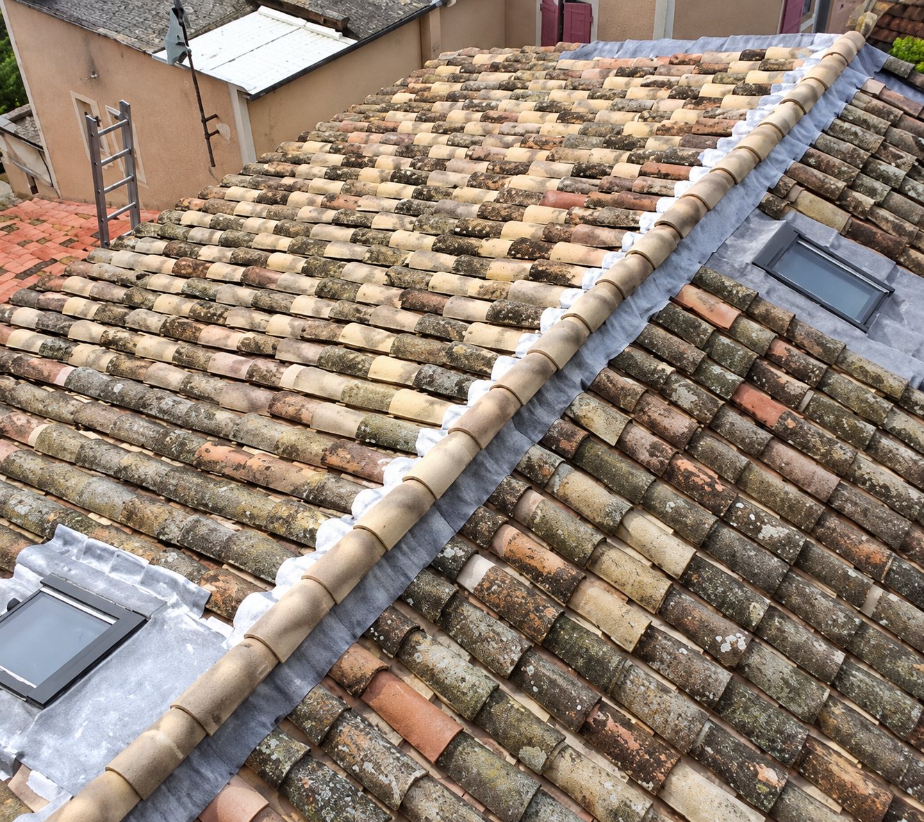 Vue en hauteur d'une toiture en vieilles tuiles canal avec deux fenêtres de toit, un faîtage et des raccords en zinc, près d'une échelle.
