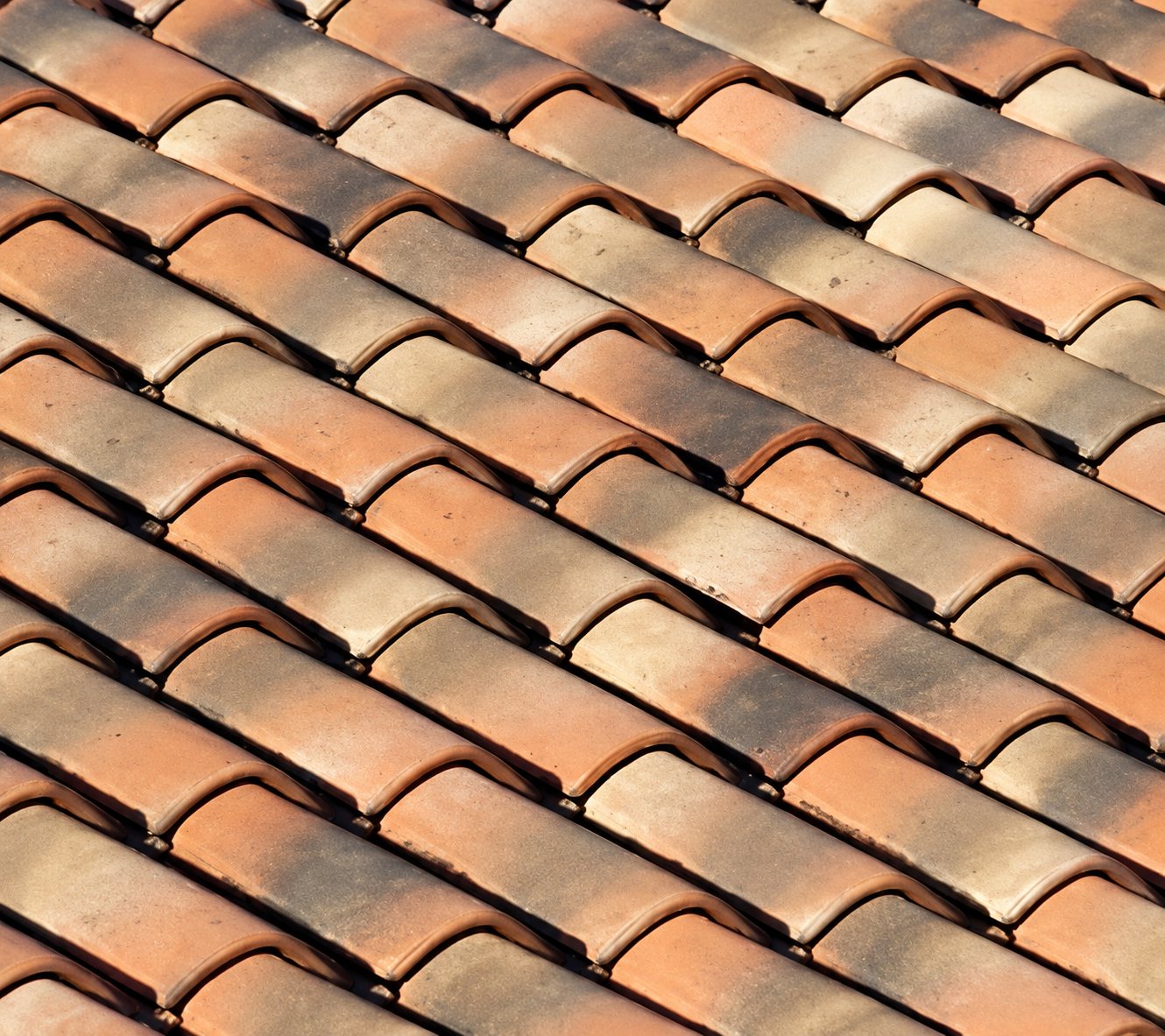 Gros plan sur une toiture de tuiles en terre cuite aux nuances orangées, beiges et brunes, disposées en rangées régulières.